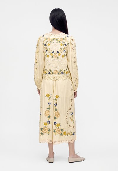Woman with long black hair wearing a cream dress with yellow and blue floral embroidery and lace details, standing facing away on a white background.