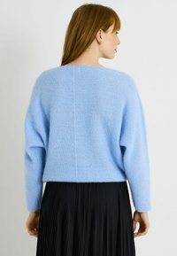 Pull en tricot bleu clair avec des épaules tombantes et des manches longues, associé à une jupe plissée noire. Le pull présente une finition texturée.