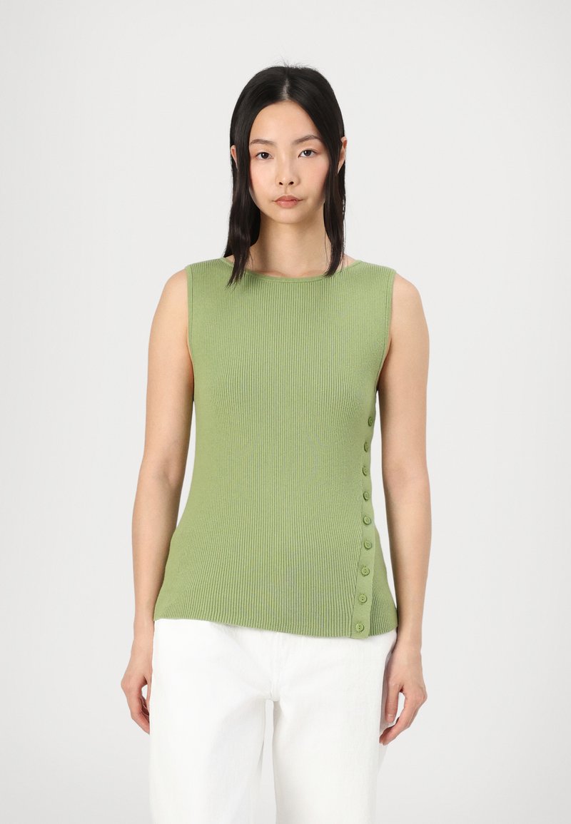 Top verde a costine senza maniche con un collo alto e una fila di bottoni lungo il lato sinistro. Abbinato a pantaloni di tessuto bianchi.