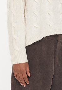 Svijetložuti pulover s pletenim uzorkom i rebrastim rubom, uparen s tamnosmešnim kordon pantalonama. Ruka odmara na rubu pulovera.