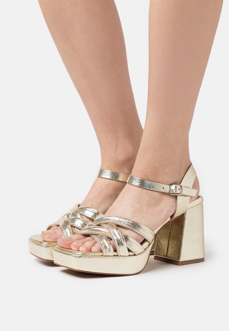 Sandales plateformes métalliques dorées avec un talon épais, dotées de brides croisées et d'une boucle à la cheville. Texture lisse et finition brillante.