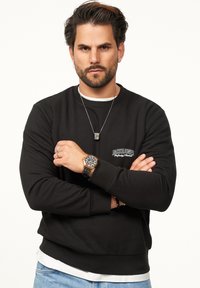 Black sweatshirt with white logo, long sleeves, and crew neck. Worn over a white t-shirt, paired with blue denim jeans and a watch.