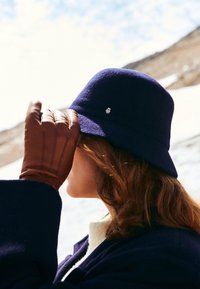 Marineblauer Woll-Bucket-Hat mit kleinem Logo an der Seite, kombiniert mit braunen Lederhandschuhen mit gestickten Details.