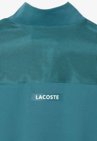 T-shirt de sport teal présentant une section supérieure en maille, une section inférieure unie et un logo Lacoste apposé sur la partie inférieure. Texture lisse dans l'ensemble.