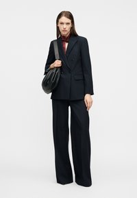 Γυναίκα που στέκεται με μαύρο σακάκι διπλής σειράς κουμπιών και παντελόνι με φαρδύ πόδι, μπορντό μεταξωτό μαντήλι και κρατώντας μια μαύρη δερμάτινη τσάντα ώμου.