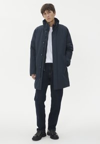 Jūras zilas krāsas parka jaka ar augstu apkakli, priekšējo rāvējslēdzēju un pogām. Uzvilkta virs baltas T-kreklu un tumšām džinsām, ar sānu kabatām.