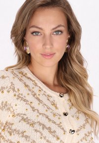 Cardigan de punto beige con un patrón de cuentas doradas, botones en forma de corazón y acentos texturizados. La modelo lleva pendientes grandes de oro y púrpura.