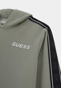 Tamsiai žalia su kapuce džemperis su tekstūruota apdaila. Yra juodos šoninės juostos su logotipo raštu ir sidabriniais užrašais 'GUESS'.