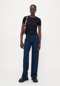 Femme avec des cheveux tressés portant un haut noir à manches courtes, un jean bleu foncé, des chaussures noires chunky et un sac à bandoulière bordeaux, se tenant devant un fond blanc.