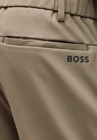 Κοντινό πλάνο στην τσέπη μπεζ παντελονιού με θηλιά για ζώνη και το κείμενο "BOSS" τυπωμένο κοντά στη ραφή της τσέπης.