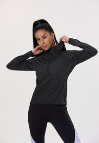 Schwarzer sportlicher Pullover mit hohem Kragen, halbem Reißverschluss und strukturiertem Stoff. Kombiniert mit schwarzen Leggings mit hellblauen Akzenten.