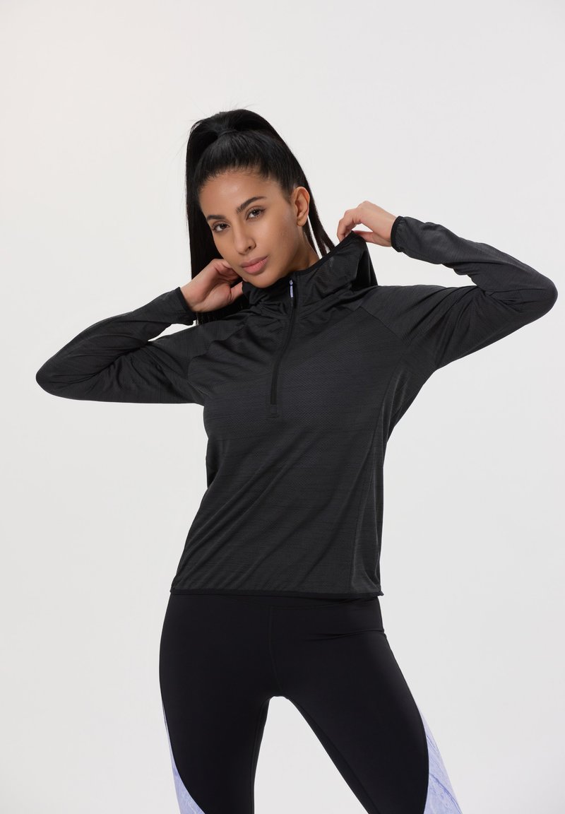 Schwarzer sportlicher Pullover mit hohem Kragen, halbem Reißverschluss und strukturiertem Stoff. Kombiniert mit schwarzen Leggings mit hellblauen Akzenten.
