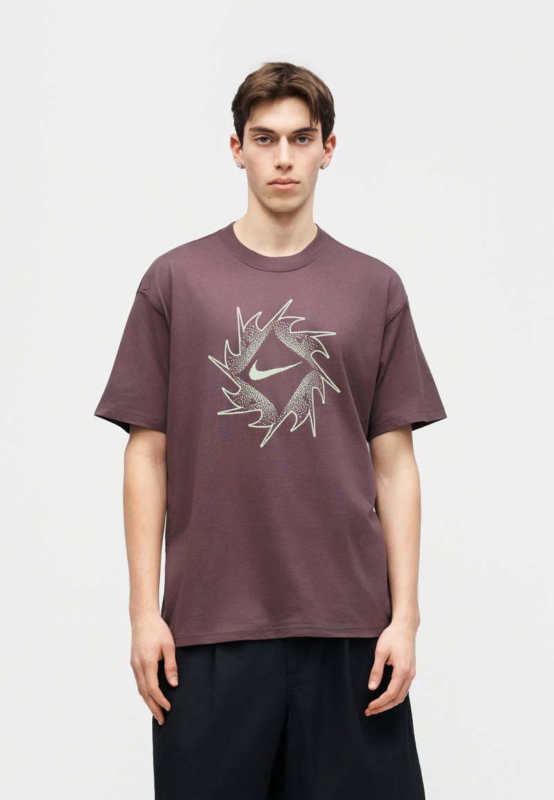 Junger Mann trägt ein übergroßes lila T-Shirt mit einem weißen abstrakten runden Nike-Logo und steht vor einem schlichten hellen Hintergrund.