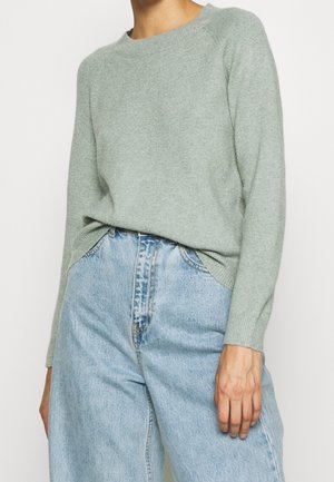 Femme portant un pull en maille vert clair rentré dans un jean en denim bleu clair à taille haute, debout devant un fond uni.
