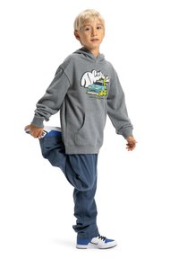 Sivá mikina s grafickým potlačou, modré nohavice a biele tenisky s modrými akcentmi. Oblečenie má voľný strih a mäkké materiály.