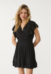 Robe noire avec un design en portefeuille, manches courtes évasées, taille smockée et ourlet à volants superposés. Fabriquée dans un tissu léger et texturé.