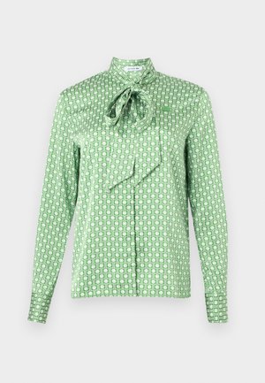 Blouse verte à manches longues avec motif géométrique blanc, col haut et nœud noué au niveau du cou.