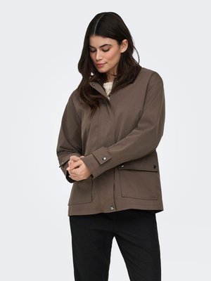 Mujer con cabello oscuro ajustándose el puño de una chaqueta marrón con botones a presión, vistiendo pantalones negros, sobre un fondo claro y liso.