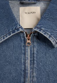 Denimjakke med krave og kobberlynlås, delvist lukket, og hvid stoflabel med teksten "SCALPERS" indeni halsudskæringen.