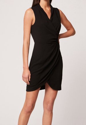 Schwarzes ärmelloses Wickelkleid mit V-Ausschnitt und drapiertem Vorderteil, getragen von einer Person, die mit einer Hand in der Hüfte vor einem einfarbigen Hintergrund steht.