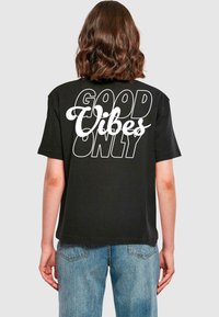 T-shirt noir en coton à manches courtes, avec un texte blanc indiquant "GOOD VIBES ONLY" imprimé au dos dans différentes polices.