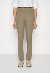 Calças numa cor azeitona clara, com corte ajustado e uma textura suave, apresentando costuras verticais. Usadas com uma camisa branca de botões e sapatos bege.