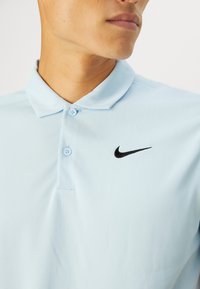 Svetlomodrá polo košeľa vyrobená z hladkého materiálu, s čiernym logom Nike swoosh a dvoma modrými gombíkmi pri golieri.