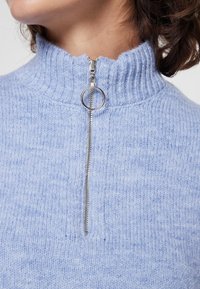 Vaaleansininen vetoketjullinen villapaita, jossa on korkea ribbitoitu kaulus. Varustettu hopeisella vetoketjulla, jossa on pyöreä vedin ja teksturoidulla neulekuviolla.