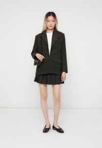 Vrouw in een oversized blazer met een donkere ruit, een wit topje, een zwarte plooorrok, die een kleine donkere tas vasthoudt en burgundy ballerina's draagt, staand.