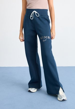Persoon draagt marineblauwe Nike joggingbroek met witte trekkoorden en logo, gecombineerd met witte en grijze sneakers.