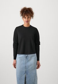 Črna pulover s krožnim ovratnikom in dolgimi rokavi, sproščenega kroja, v kombinaciji s svetlim denim krilom z odprtino spredaj in kontrastnim šivom.