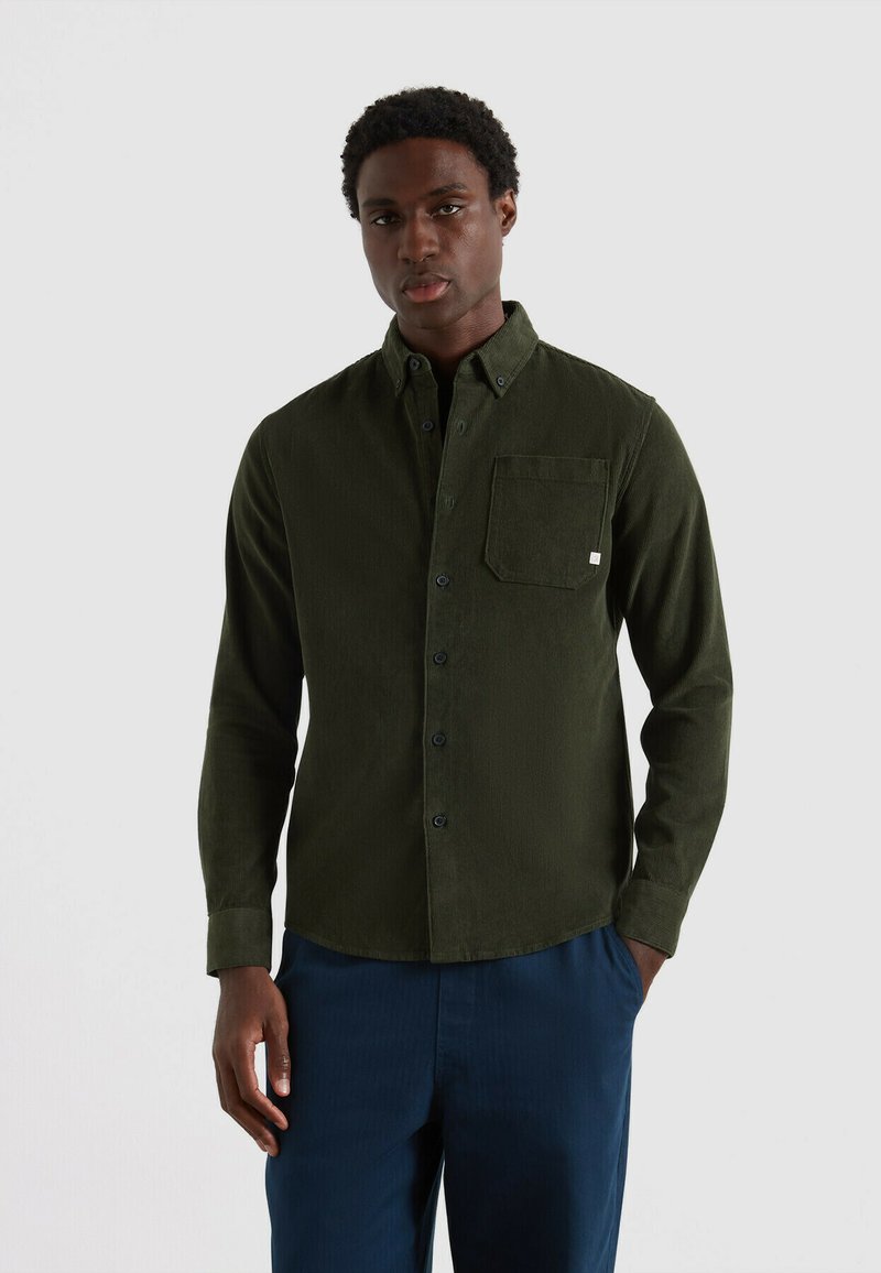 Camicia in corduroy verde scuro con colletto a bottoni, taschino frontale e bottoni neri, abbinata a pantaloni blu navy.
