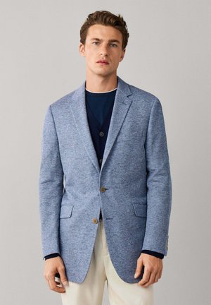 Joven vestido con una chaqueta azul claro texturizada sobre un jersey azul marino y pantalones blancos, de pie frente a un fondo liso gris claro.