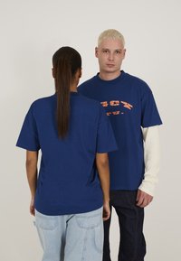 Marineblå oversized t-shirts, en med orange grafisk tekst, blødt bomuldsstof, afslappet pasform, korte ærmer og rund hals.