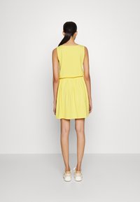 Ärmlocklös gul klänning med en tight topp och elastisk midja, flödande kjol. Modellen bär ljusa skor. Enfärgad bakgrund.