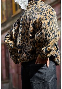 Personne portant une veste en fausse fourrure à imprimé léopard et un jean en denim foncé, les mains dans les poches arrière, standing en extérieur près d'un bâtiment.