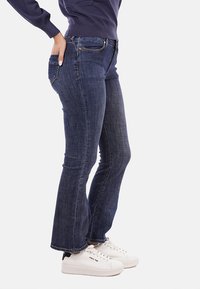 Jeans a zampa d'elefante in denim blu scuro con vita media, dotati di cinque tasche e dettagli di cucitura sottili, indossati con sneakers bianche.