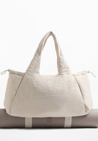 Beige quiltet totebag med brede håndtag, der hviler på en rullet grå yogamatte sikret med to beige stropper.