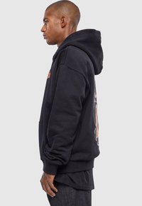 Hanorac negru din bumbac, cu buzunar tip pouch, manșete ribbate și un design grafic îndrăzneț pe spate. Croială oversized cu glugă ajustabilă prin șnur.