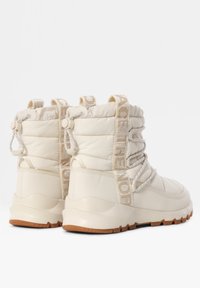 Botas acolchadas color crema con una parte superior sintética texturizada, que incluyen un sistema de cordones y una correa con un logo prominente en la parte trasera. Suela de goma para tracción.