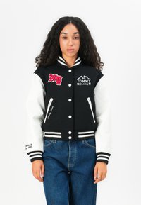 Giacca varsity nera con maniche in pelle bianca, caratterizzata da testo e toppe ricamate, polsini e orlo a coste e tasche laterali.