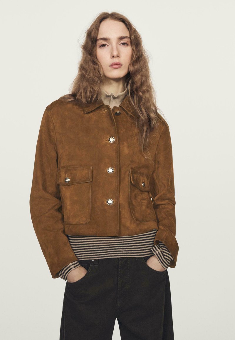 Donna con lunghi capelli ondulati che indossa una giacca in suede marrone sopra una maglietta a righe e pantaloni neri, in piedi con le mani nelle tasche su uno sfondo neutro.