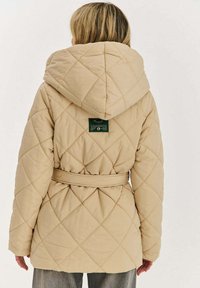 Béžová prošívaná péřová bunda s kapucí, páskem v pase a zelenou štítkem s pokyny na zadní straně, zdobená diamantovým vzorem po celém povrchu.