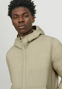 Veste légère beige avec capuche et finition texturée, dotée d'une fermeture éclair sur le devant et d'un logo brodé "Originals Studio" sur l'ourlet.
