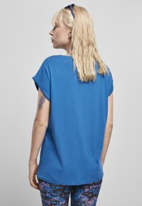 Kék pamut póló felgöngyölt ujjakkal, laza szabással és kerek nyakkivágással, lila és kék árnyalatú mintás leggingsszel viselve.