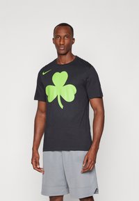 Camiseta de algodón negra con un gran diseño de trébol fluorescente verde en el frente, complementada por un pequeño logo de Nike en el hombro.