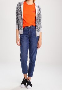 Sudadera con estampado de leopardo gris, camiseta naranja brillante, vaqueros de mezclilla azul de cintura alta y zapatillas negras sin cordones con punta blanca.