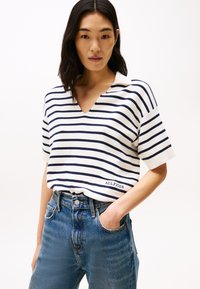 Άσπρο πουκάμισο πόλο με μπλε ρίγες και κοντά μανίκια, διαθέτοντας V-λαιμό και κεντημένο λογότυπο "HILFIGER", σε συνδυασμό με ψηλόμεσες μπλε τζιν παντελόνες.