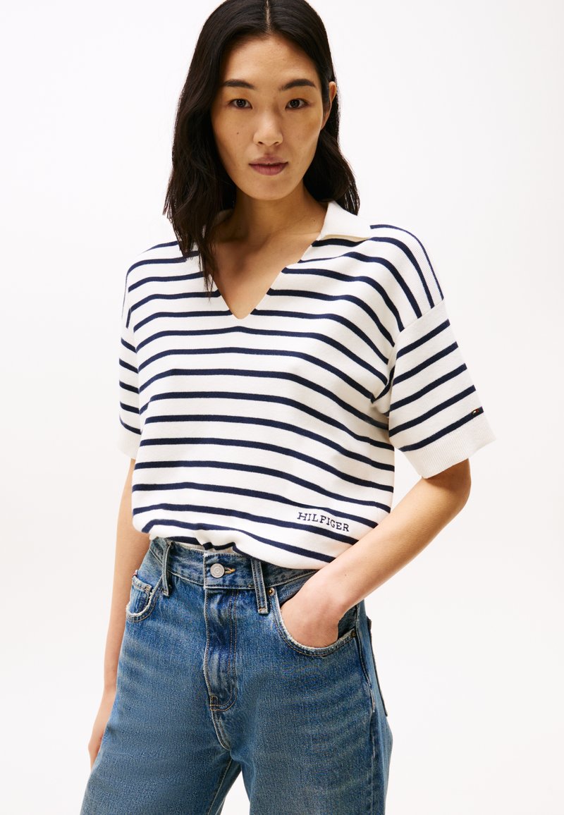 Άσπρο πουκάμισο πόλο με μπλε ρίγες και κοντά μανίκια, διαθέτοντας V-λαιμό και κεντημένο λογότυπο "HILFIGER", σε συνδυασμό με ψηλόμεσες μπλε τζιν παντελόνες.