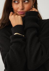 Sort strikket sweater med høj krave og ribbet tekstur. Har løse ærmer og et guld accent armbånd på håndleddet.